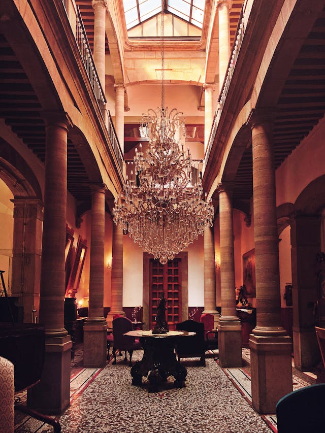 Grand crystal chandelier hanging in an elegant hotel lobby with stone columns and a glass skylight ceiling.